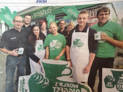 Group of volunteers in green shirts and aprons holding mugs and a banner at a World’s Biggest Coffee Morning event
