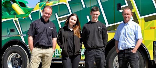 Four people standing in front of a green Manchetts truck, posing outdoors and smiling.