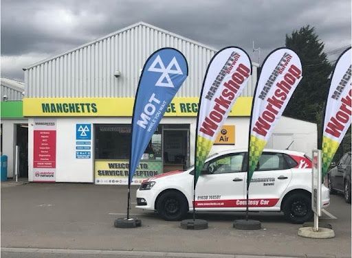 White car parked outside a Manchetts MOT garage with tall promotional flags in front of a gray building