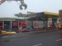Collapsed storefronts with a crumpled yellow roof; a crane claw hangs above a wet street.