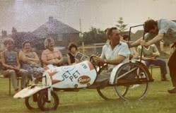 People gathered around a homemade pedal vehicle labeled “JEEP” in a village setting.