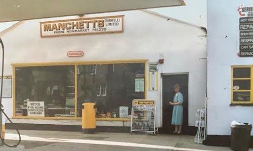 Small storefront with a yellow post, large window, and a person standing in the doorway Manchetts first garage 