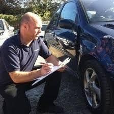 Man kneeling beside a blue car, writing on a clipboard during an inspection.