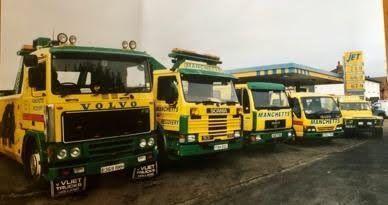 Line of colorful yellow-green older Manchetts Volvo and Mercedes tow trucks parked outdoors on a wet lot