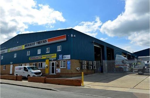Blue industrial Manchetts warehouse with parked vans and a gated entrance under a cloudy sky