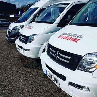 Row of white commercial vans parked outdoors, with red “Manchetts Self drive hire” signs on the fronts