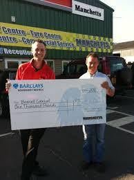 Two men holding an oversized Barclays cheque outside a Manchetts store.