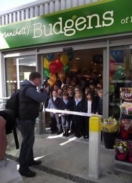 Crowd gathered outside Budgens store entrance with balloons, facing a man speaking on a phone.