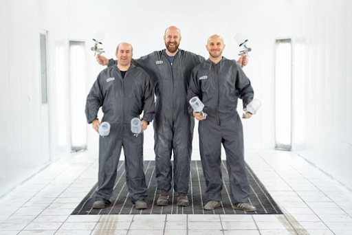 Three technicians in gray coveralls standing in a bright tiled room, each holding a tool.