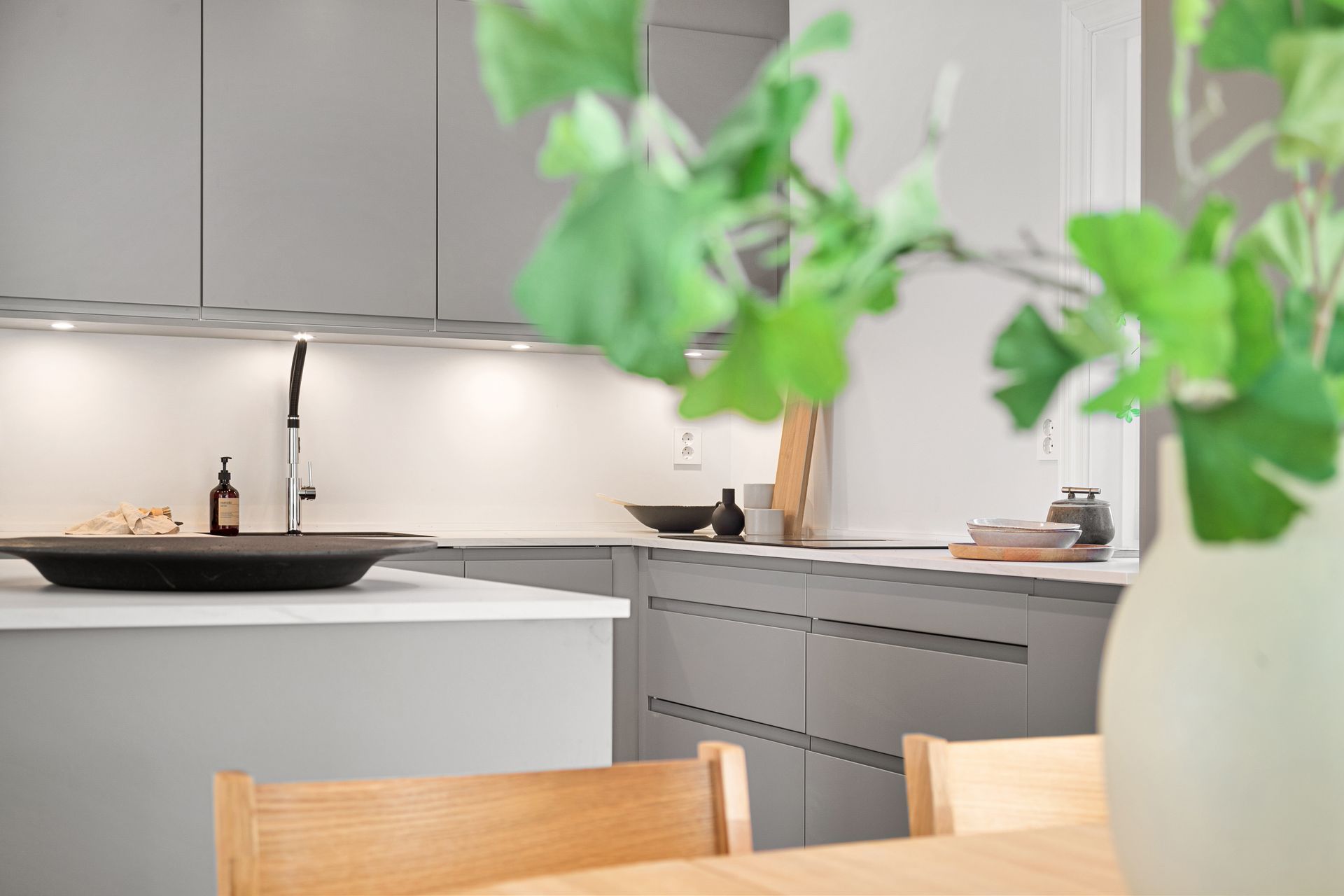 Moderne kjøkken med grå skap, hvit kjøkkenøy og uskarp grønn plante i forgrunnen