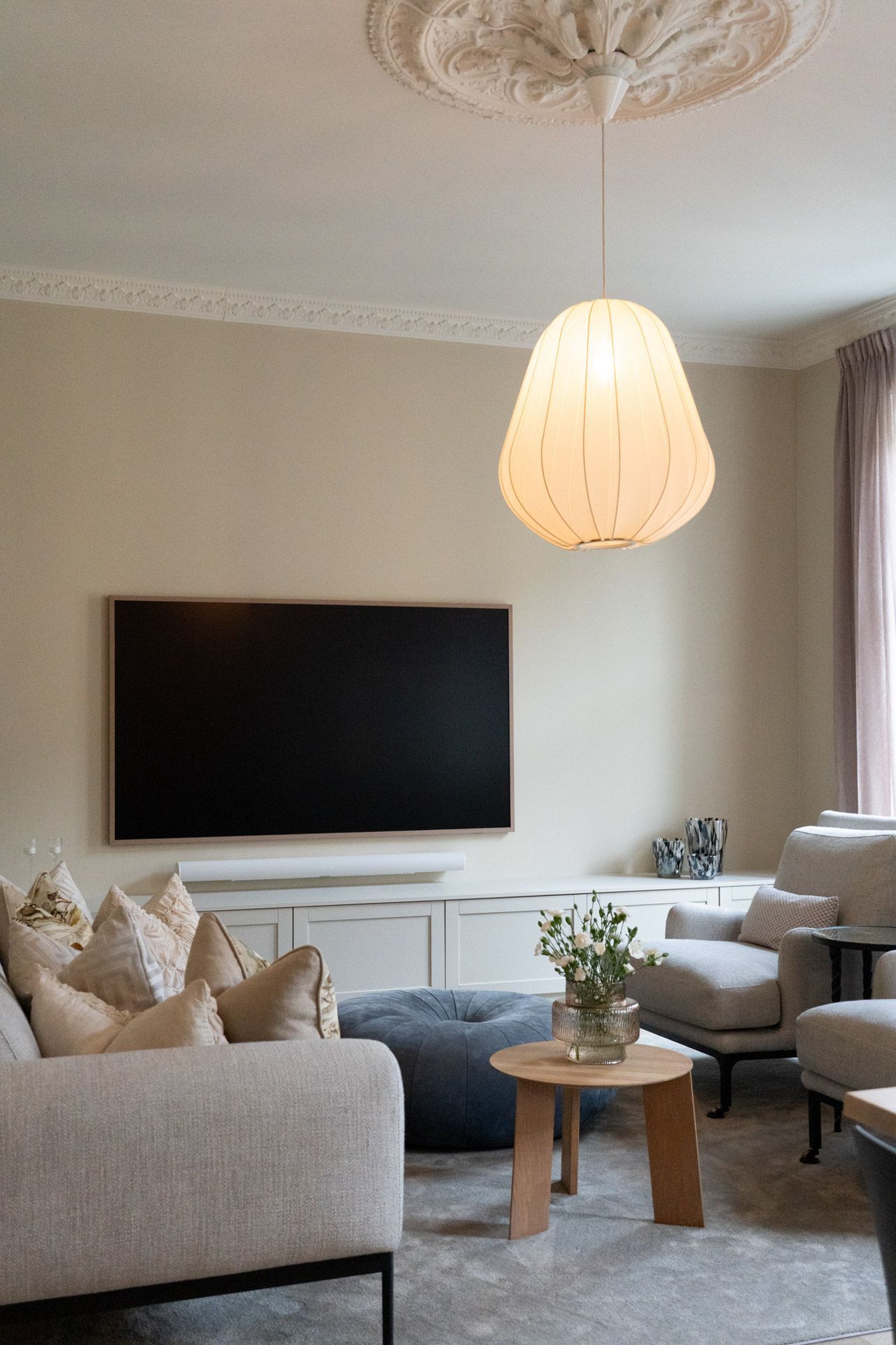 Koselig stue med vegghengt TV, kremfarget sofa, rundt trebord og hengende pendellampe.