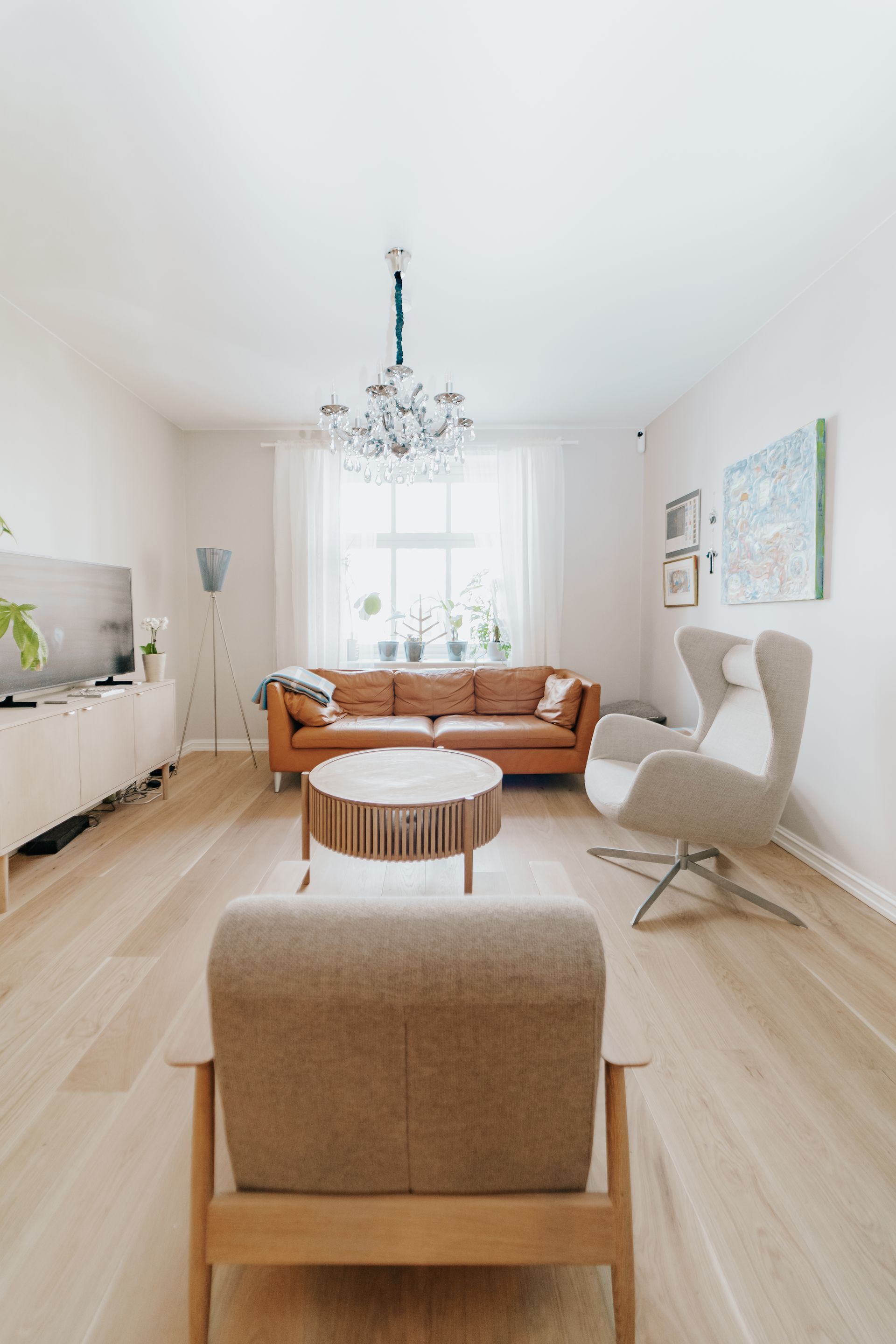 Lys minimalistisk stue med brun sofa, hvit stol, lyst tregulv og stort vindu med lysekrone