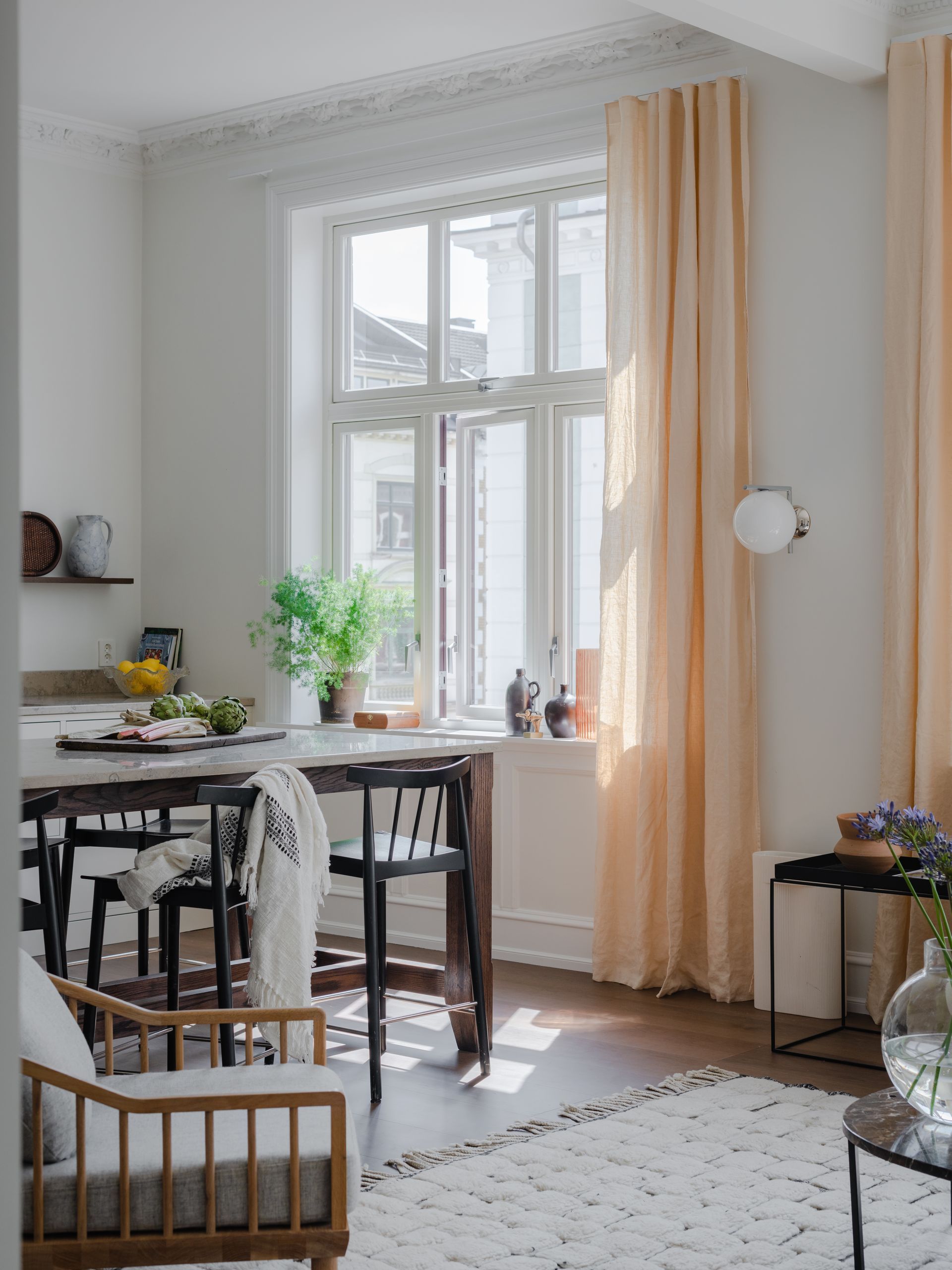 Lys spisekrok med bord, stoler, planter og et vindu innrammet av ferskenfargede gardiner i et hvitt rom