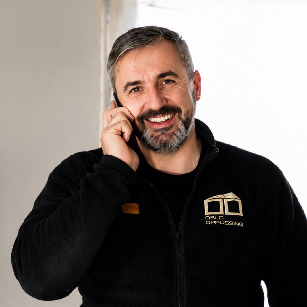 Smilende mann i svart jakke snakker i telefon mot en lysegrå bakgrunn