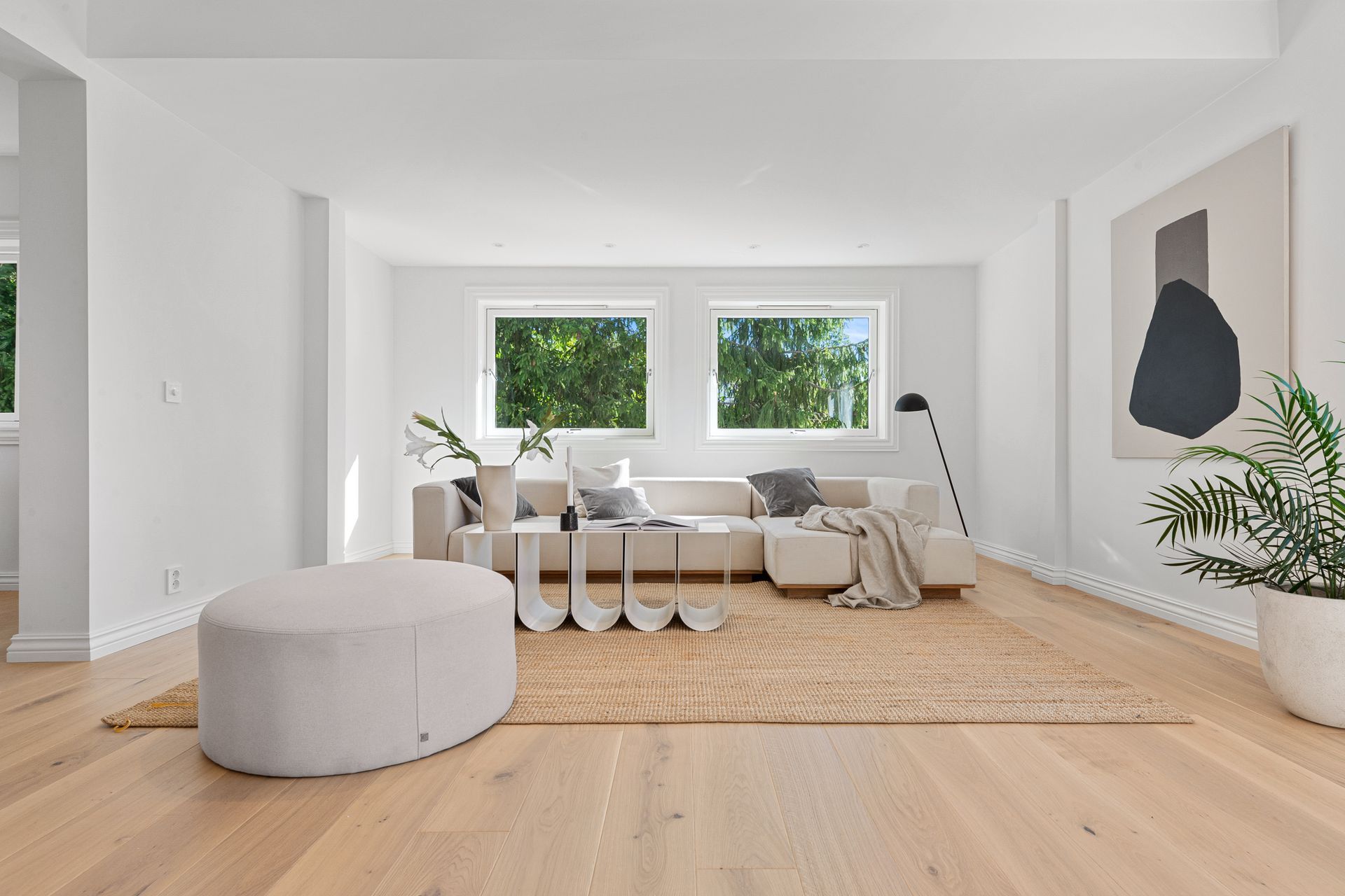 Lys minimalistisk stue med beige sofa, rund puff, vevd teppe og store vinduer.