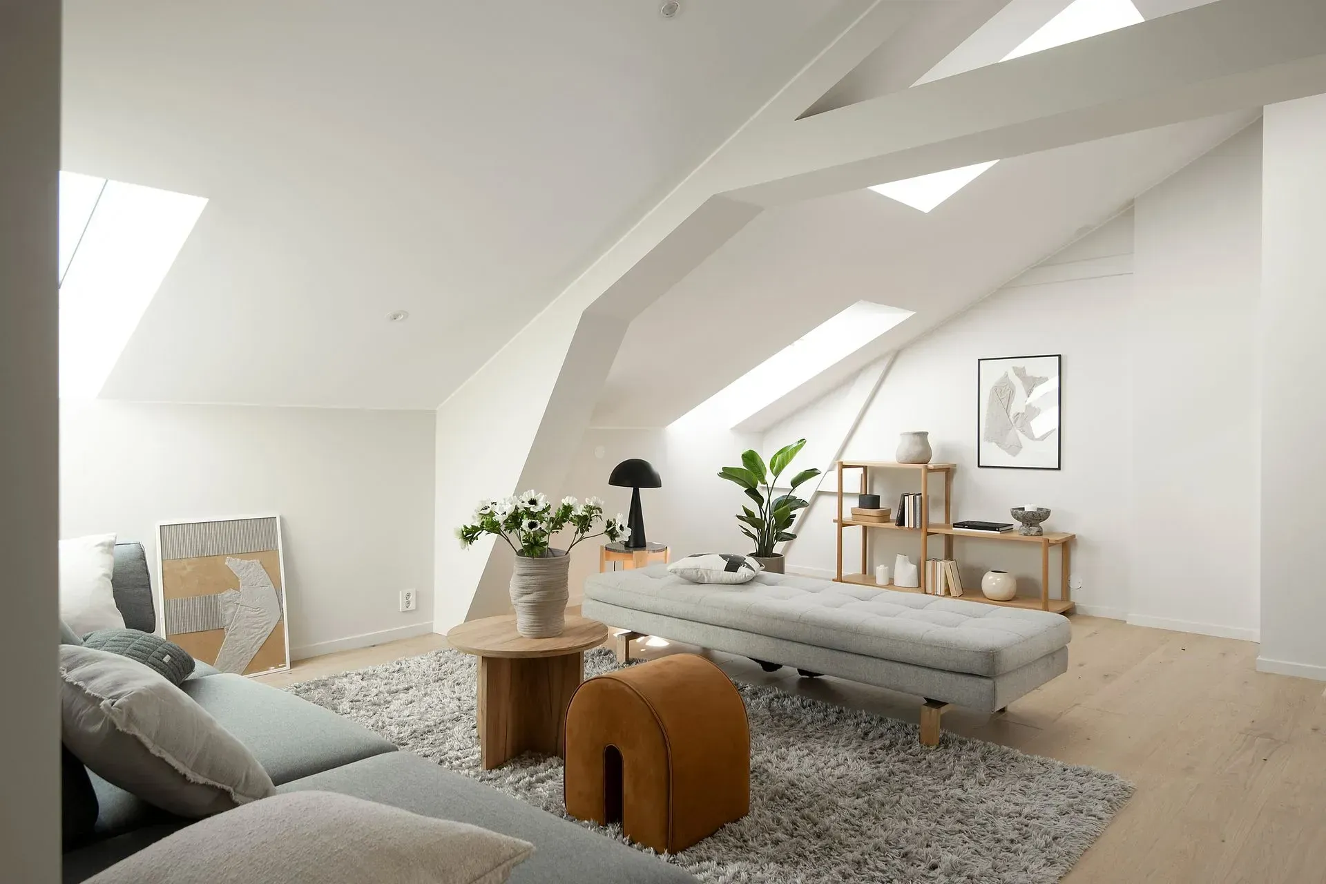 Lys loftstue med hvite bjelker, grå sofa, teppe, salongbord, planter og trehyller