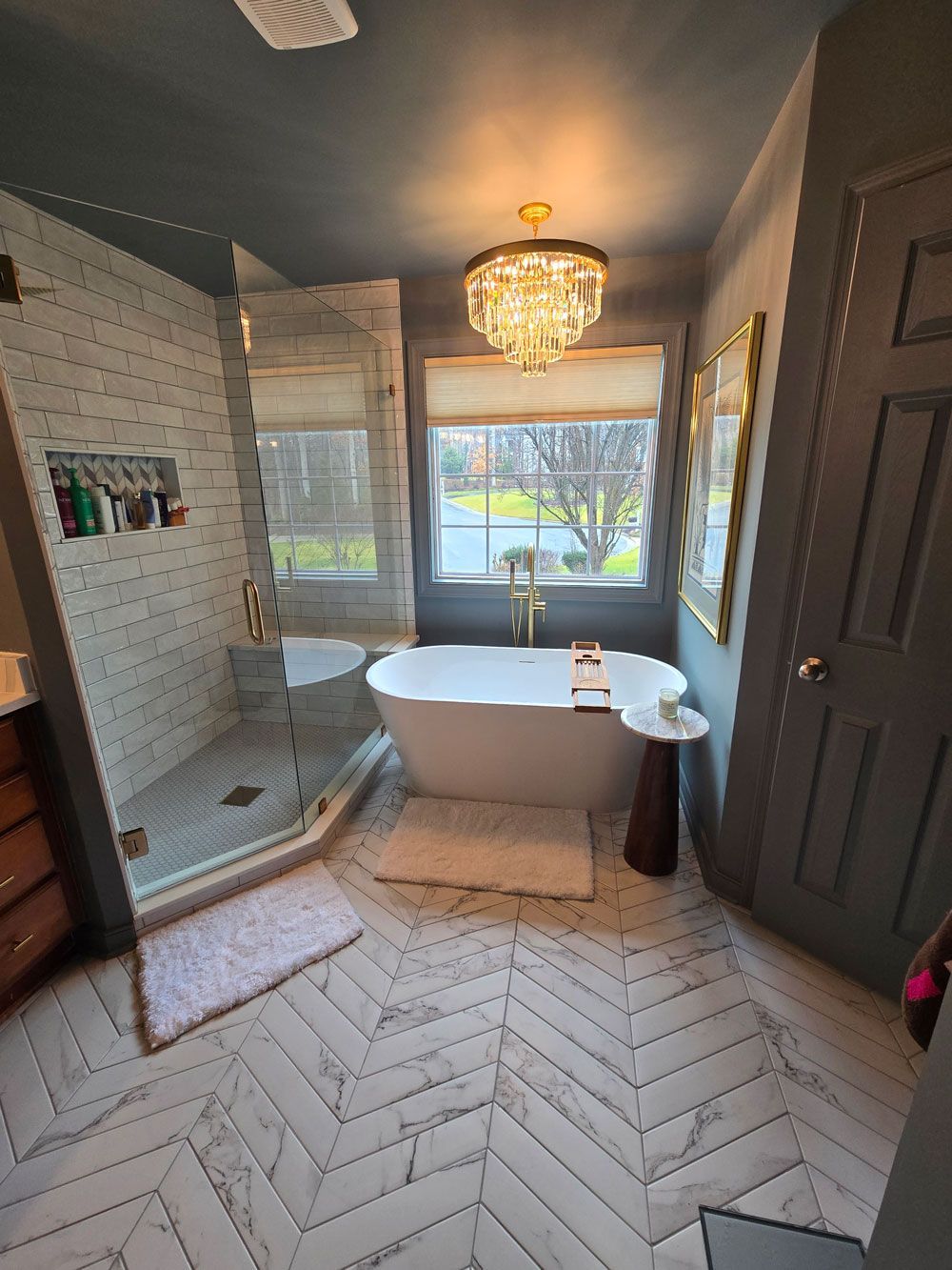 Bright bathroom with chandelier, walk-in shower, freestanding tub, and herringbone tile floor.