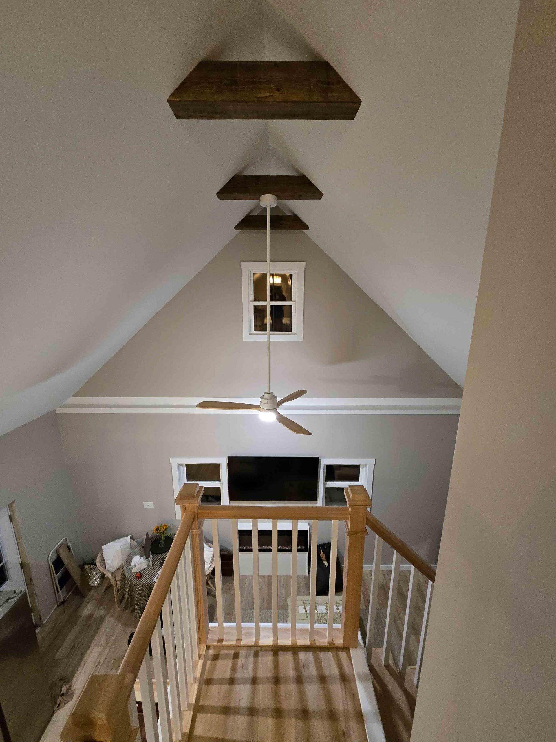 View from a staircase into a vaulted living room with a window, fan, and wooden beams.