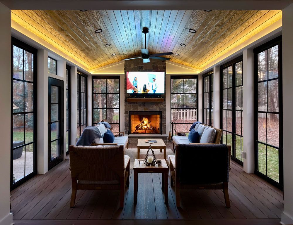 Sunroom with a fireplace, TV, and seating surrounded by large windows, with wood ceiling and floors.