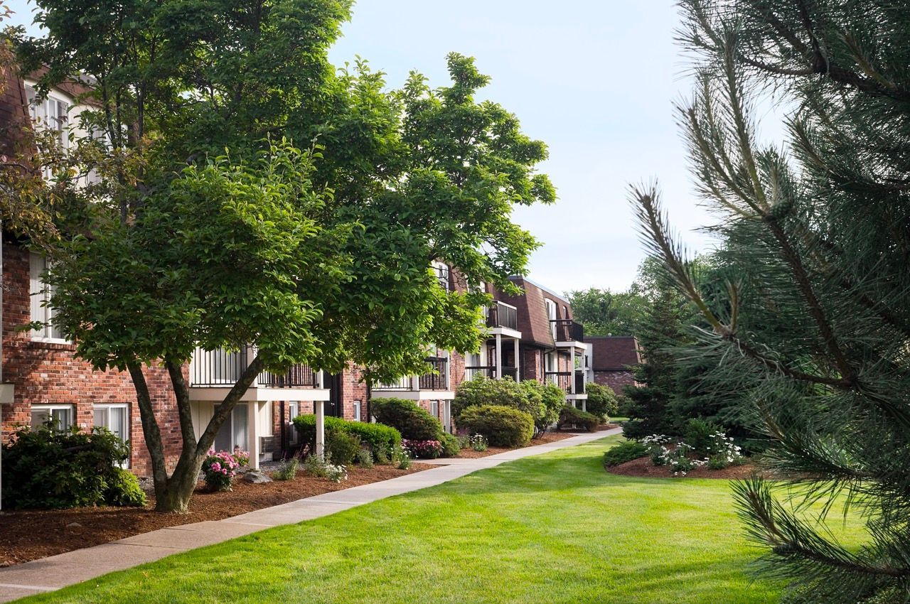 Exterior view of brick apartment buildings with a landscaped lawn, trees, and a sidewalk.