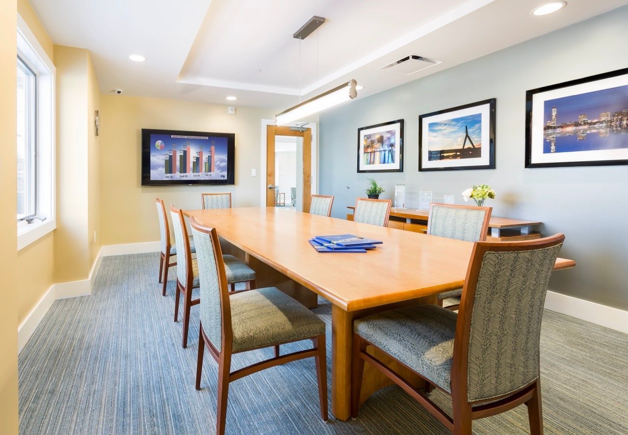 Community conference room with a long wooden table, upholstered chairs, wall art, and a wall-mounted display.
