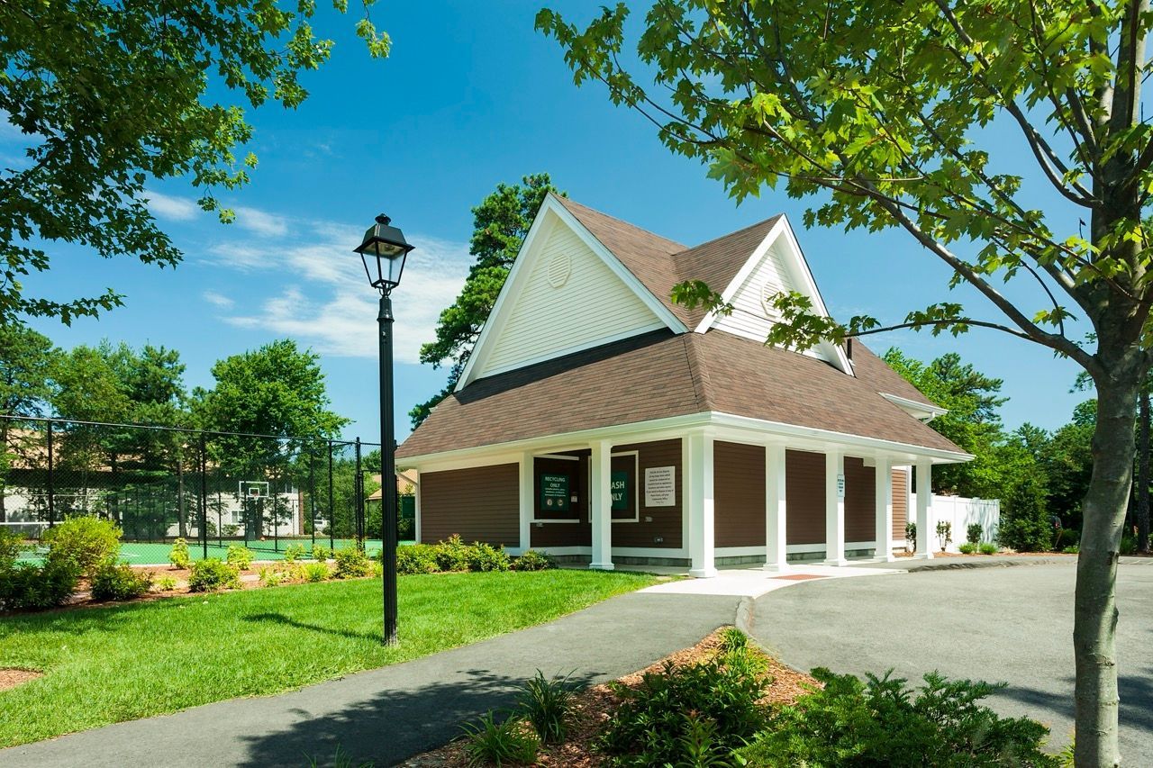 Exterior view of a brown clubhouse with white trim, surrounded by trees and a nearby tennis court.