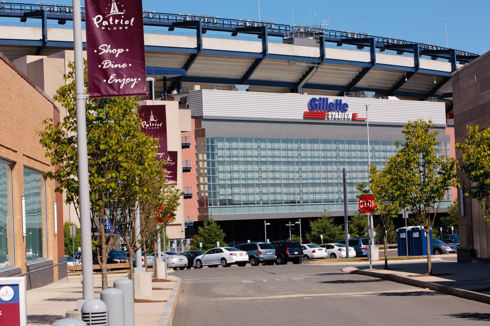 Street view of a stadium entrance with cars, trees, and a modern glass facade under a large overhang.