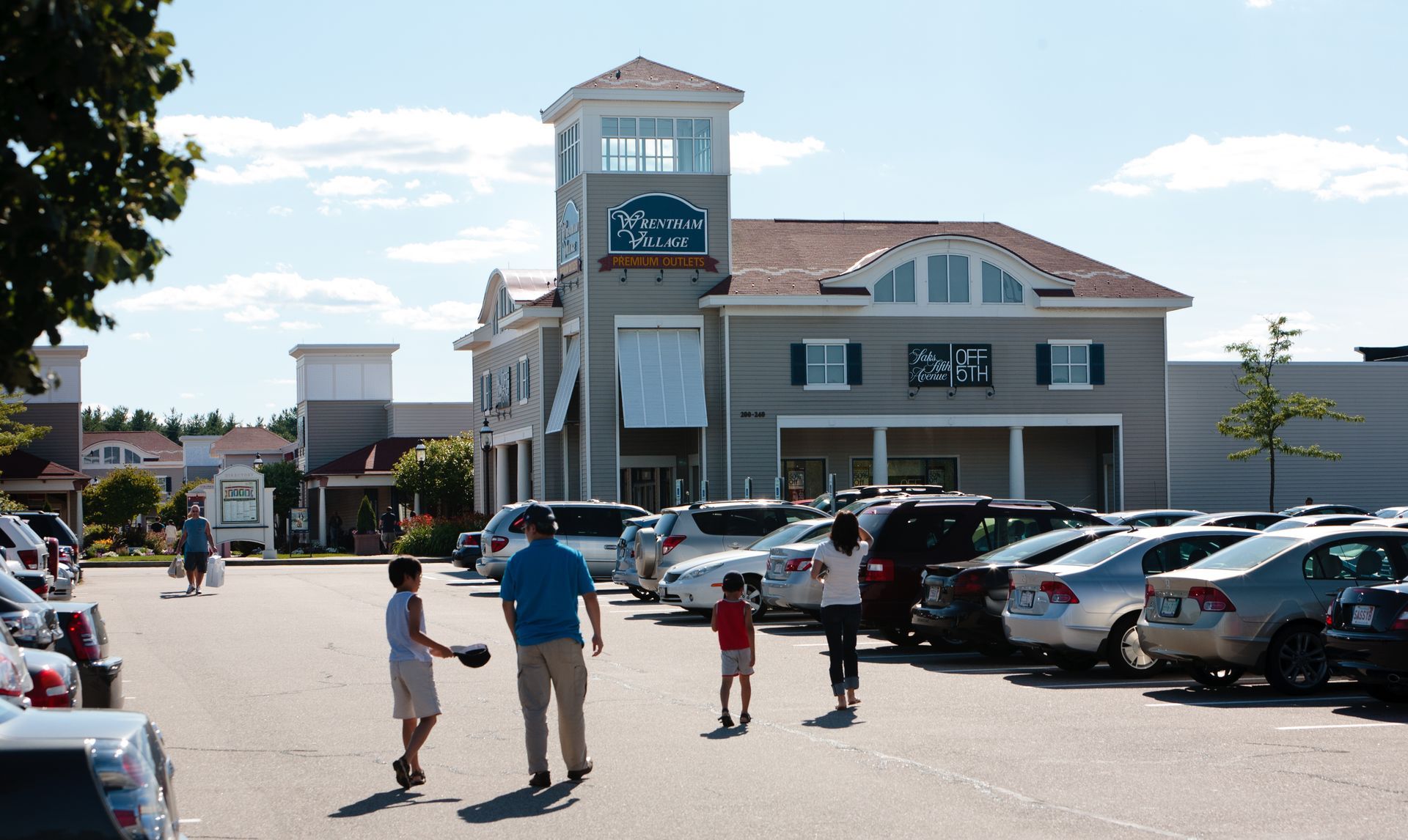 Shoppers walking toward a tan mall entrance with parked cars under a sunny sky