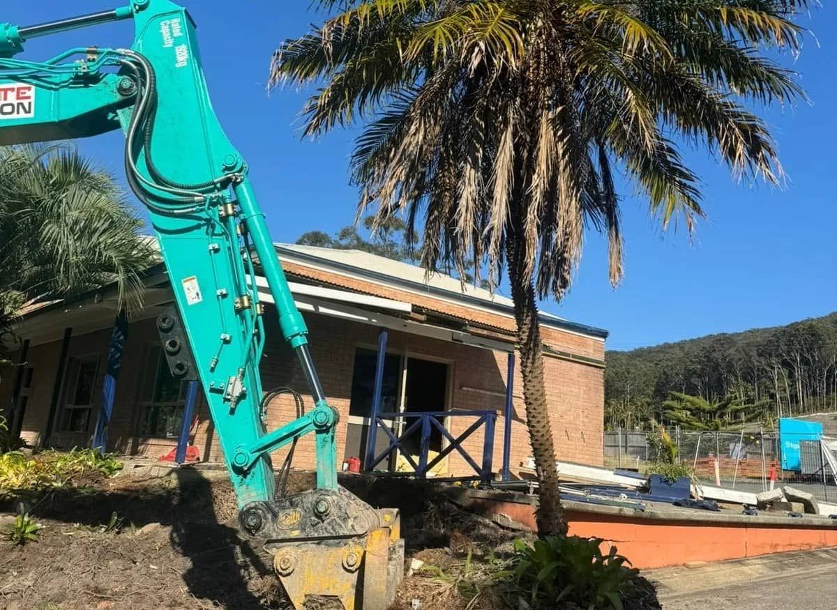 A Blue Excavator Is Parked in Front of A Building Next to A Palm Tree — Central Coast Complete Demolition & Tree Service in Buff Point, NSW