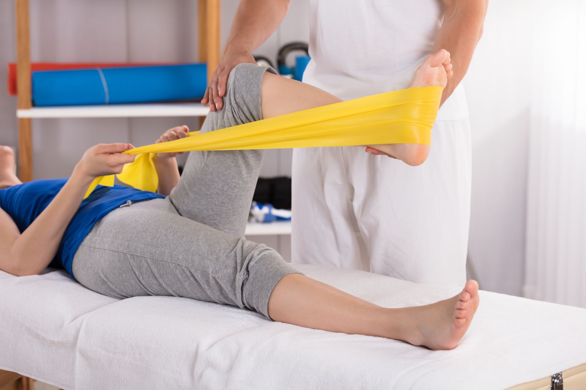 Una donna è sdraiata su un letto mentre un fisioterapista le allunga la gamba con una fascia elastica gialla.