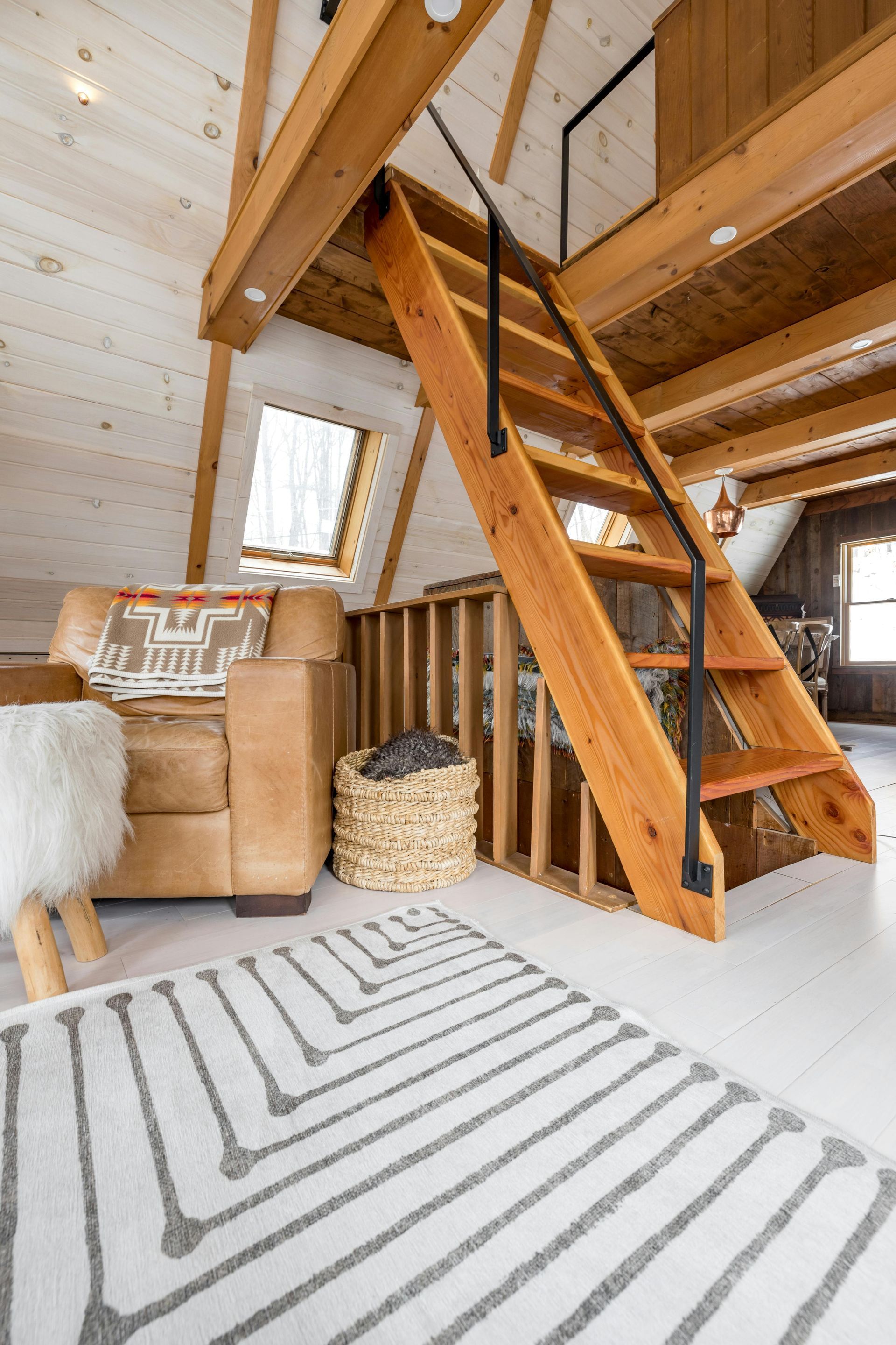 Cozy cabin interior: wooden staircase to loft, leather chair, patterned rug.