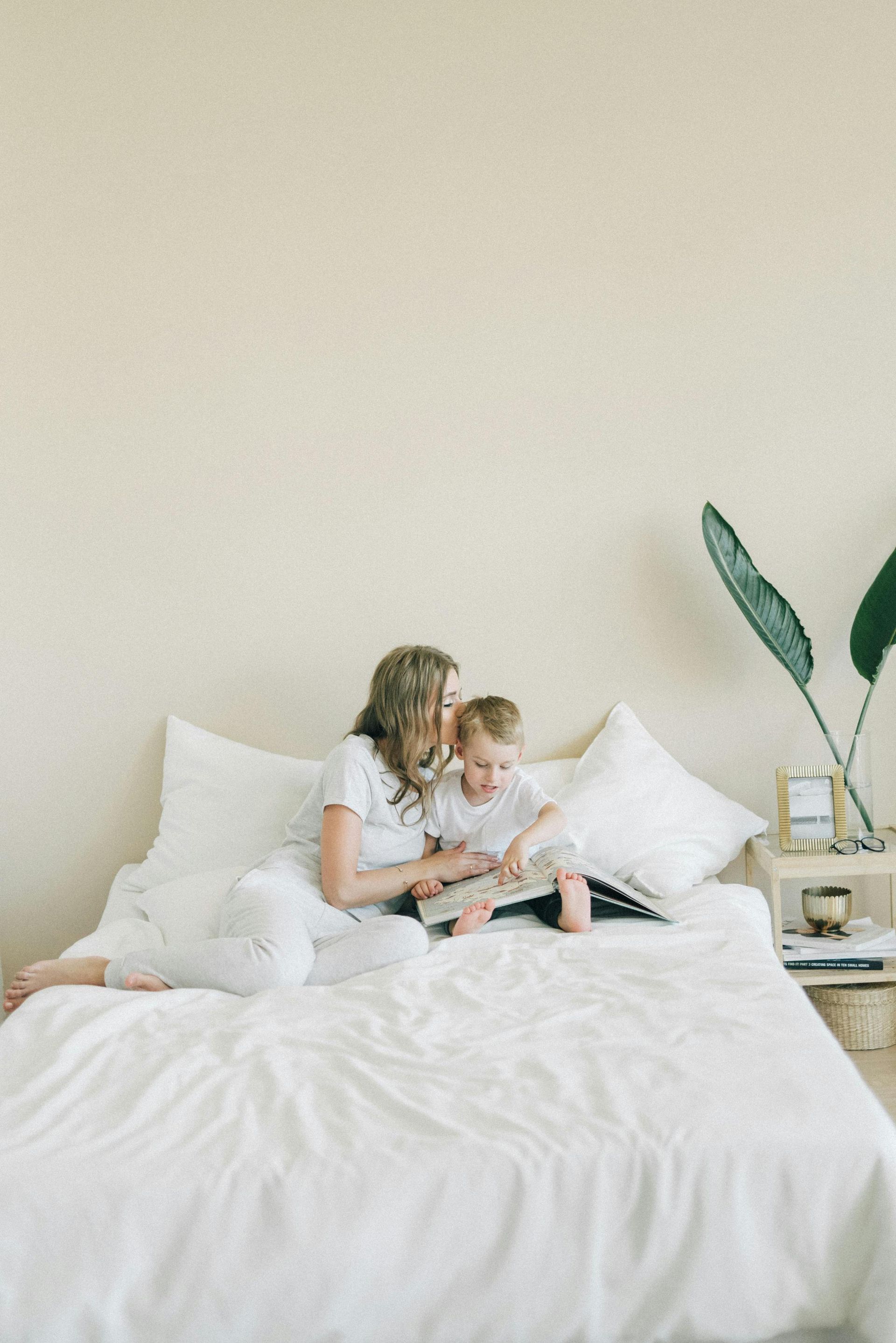 Woman and child read a book on a bed, light-colored room.