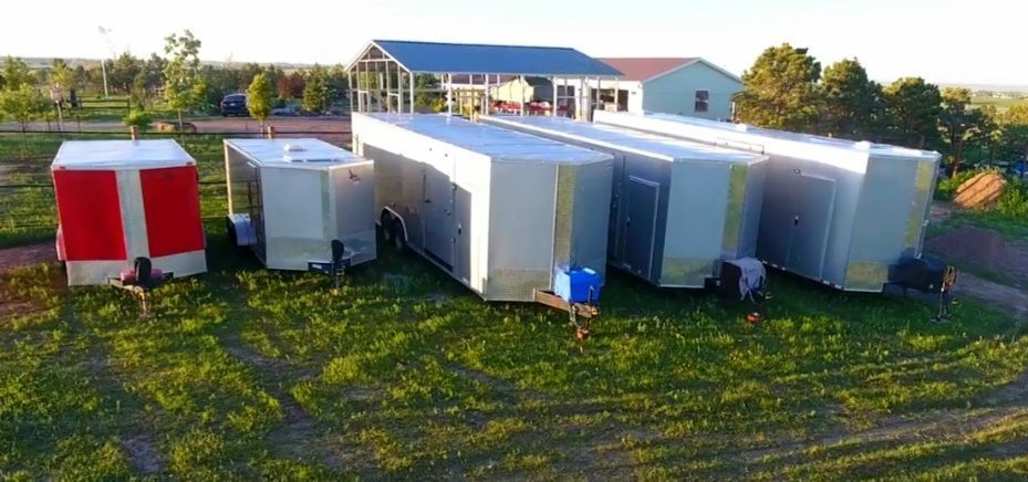 Several portable sheds and trailers on a grassy lot, including red, white, and gray units near a barn.