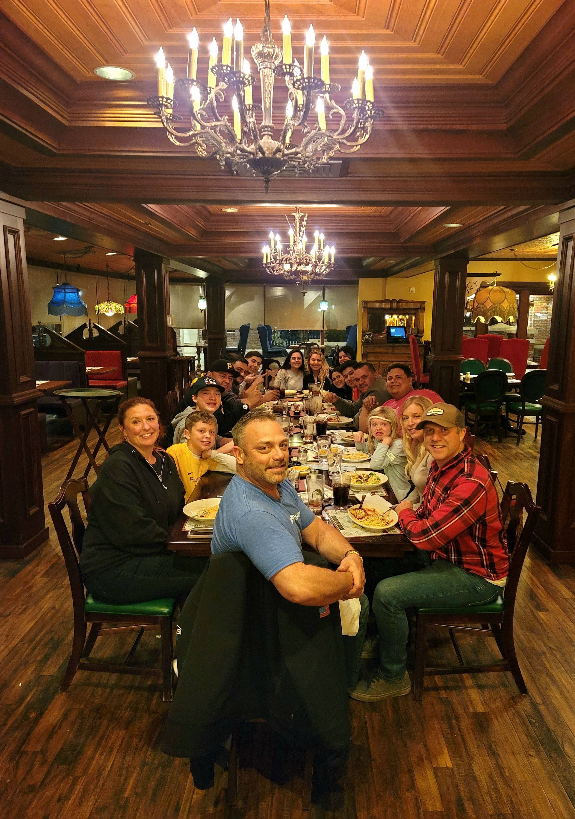 Group dining in a wood-paneled restaurant under chandeliers, seated around a long table and smiling at the camera
