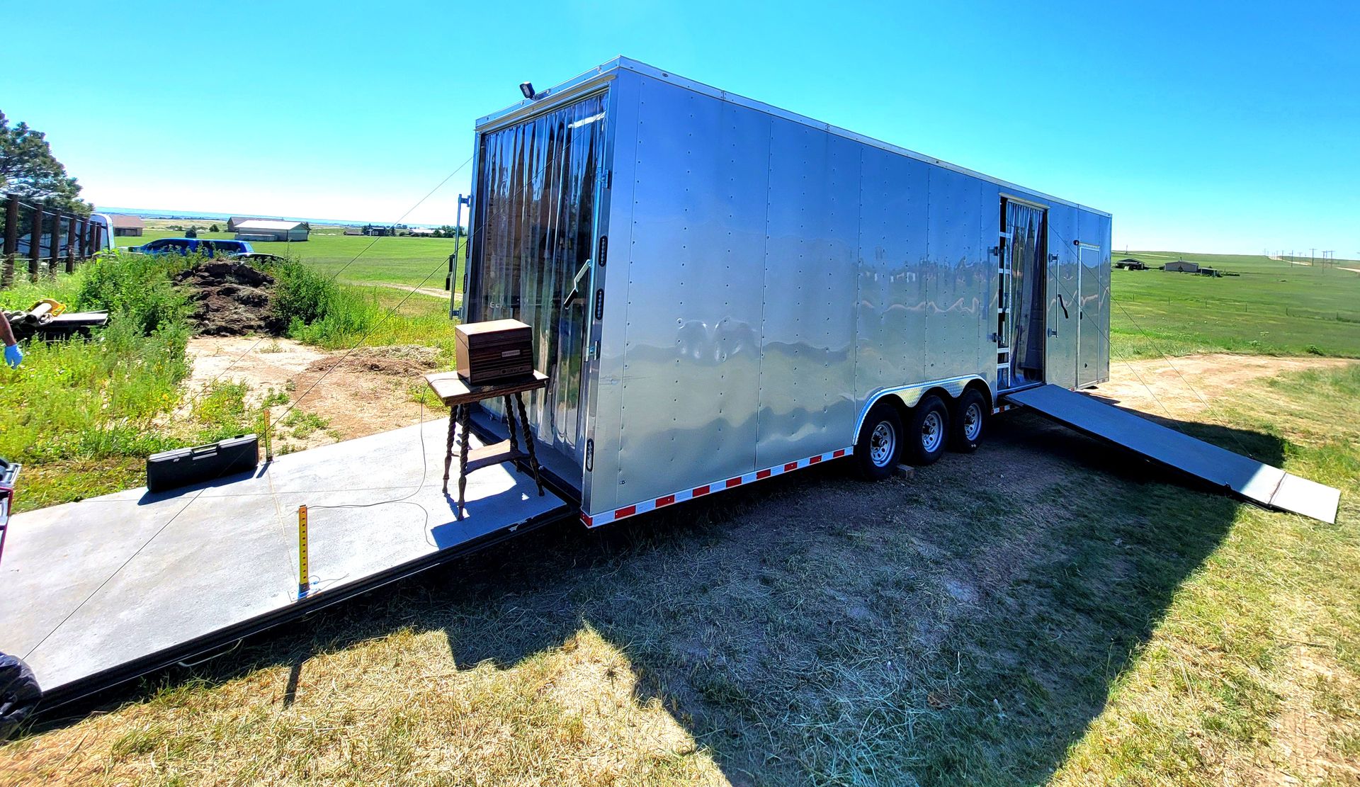 Silver enclosed trailer parked on a grassy field beside a concrete pad, with ramps extended.