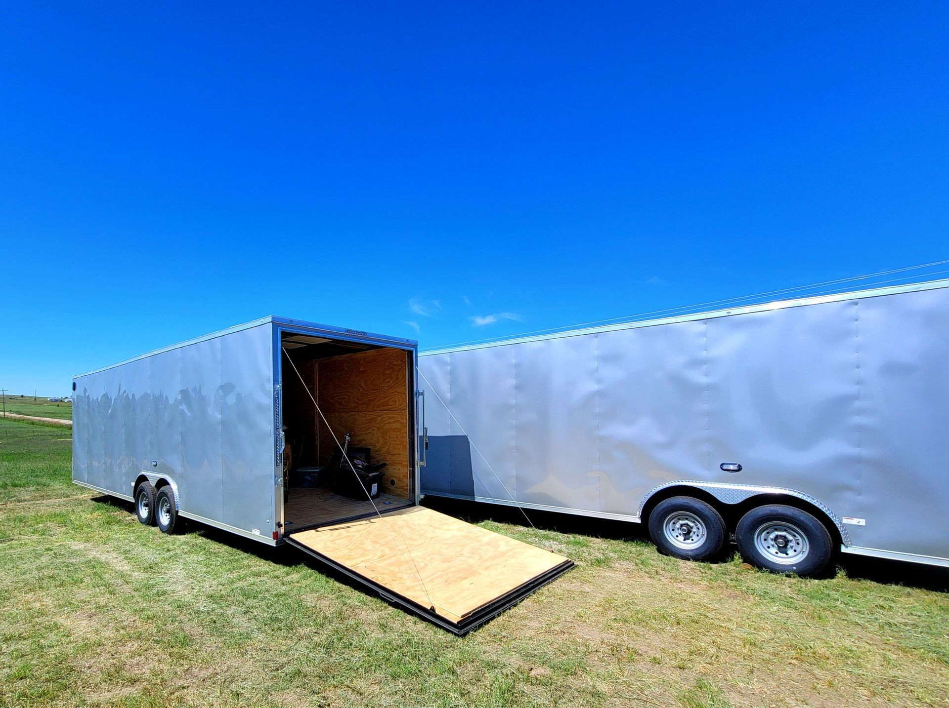 Open cargo trailer with ramp beside a closed silver trailer in a grassy field under blue sky