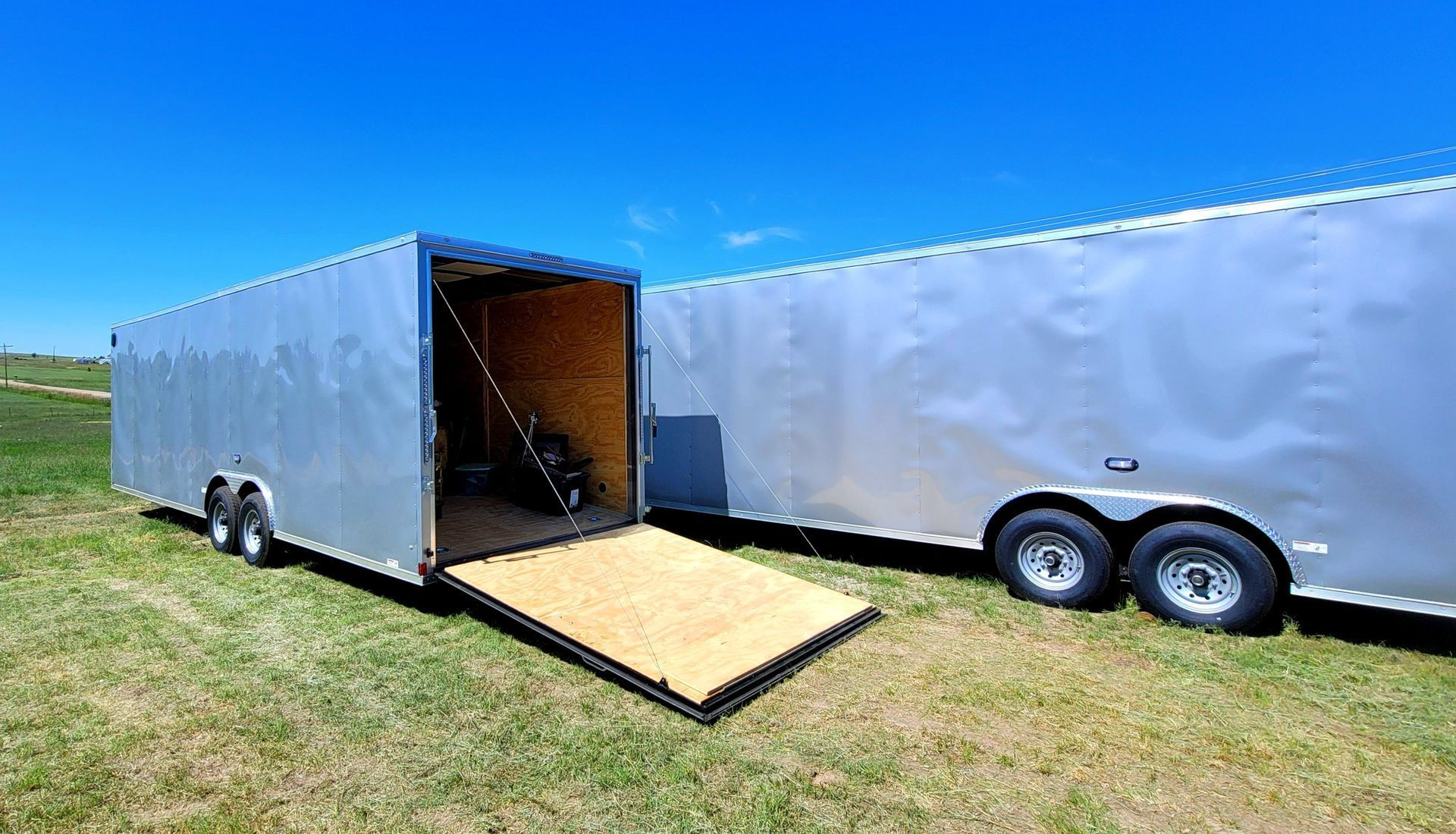 Open cargo trailer with ramp beside a large silver enclosed trailer on a grassy field under blue sky
