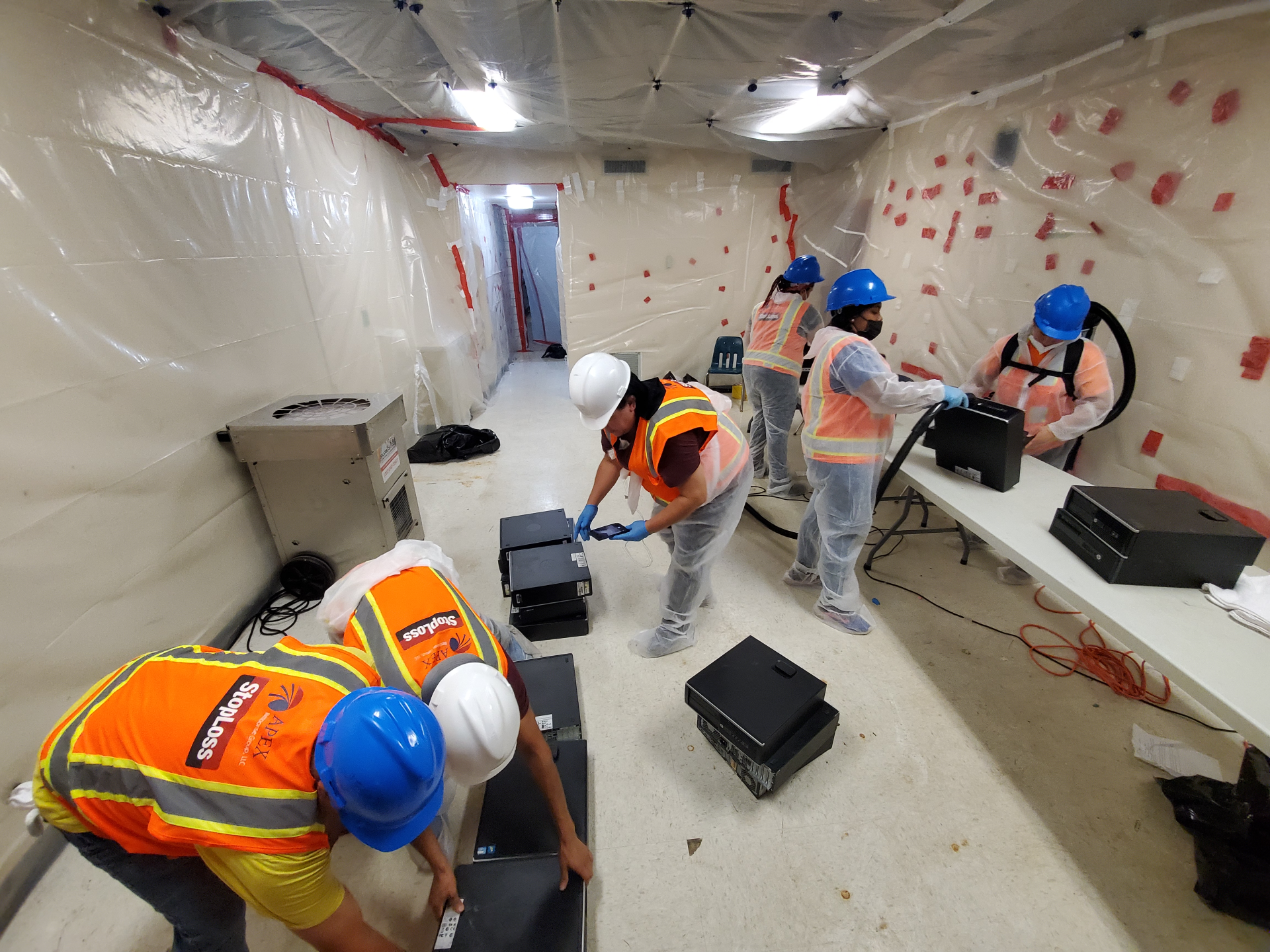 Workers in hard hats and vests installing drywall in a plastic-covered room