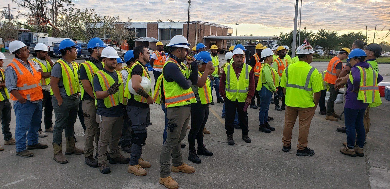 Construction workers in hard hats and safety vests gathered outdoors at a job site, listening to a briefing.