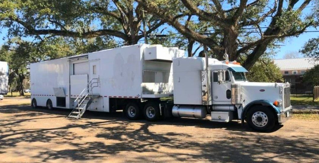 White utility truck with trailer parked under large trees on a dirt lot