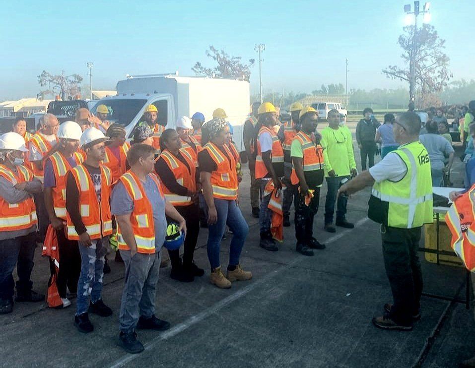 Construction crew in orange safety vests gathered for a briefing outdoors, facing a supervisor in a yellow vest.