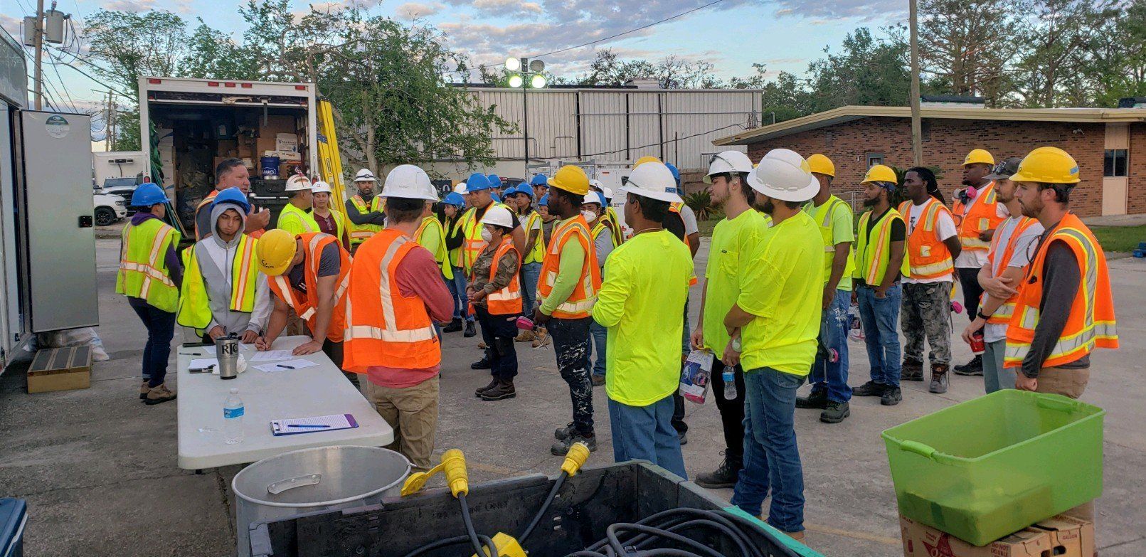 Construction crew in hard hats and safety vests gathered outdoors around equipment and materials.