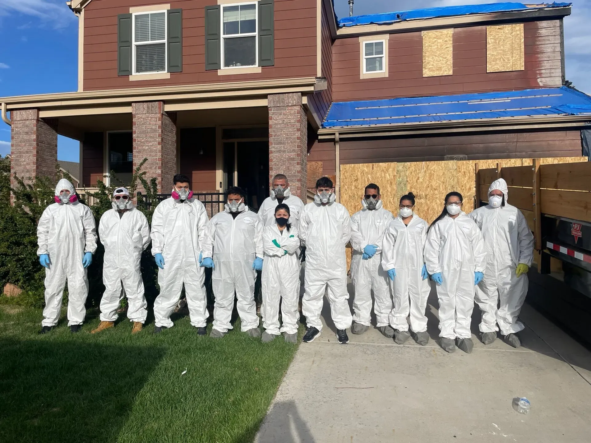 Group in white protective suits standing outside a brick house with a blue tarp roof.