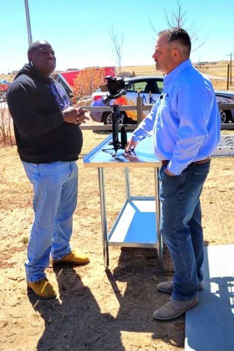 Two people talking beside a table outdoors on a sunny day, one holding a drink behind a microphone setup