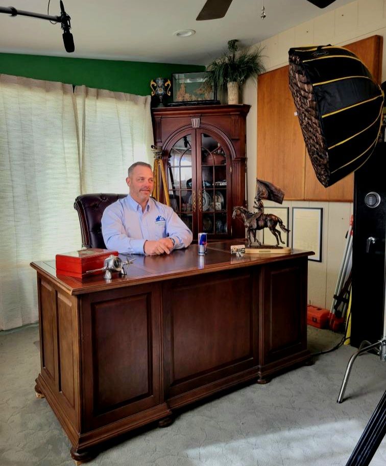 Man seated at a wooden desk in a studio with lighting equipment and a large cabinet behind him
