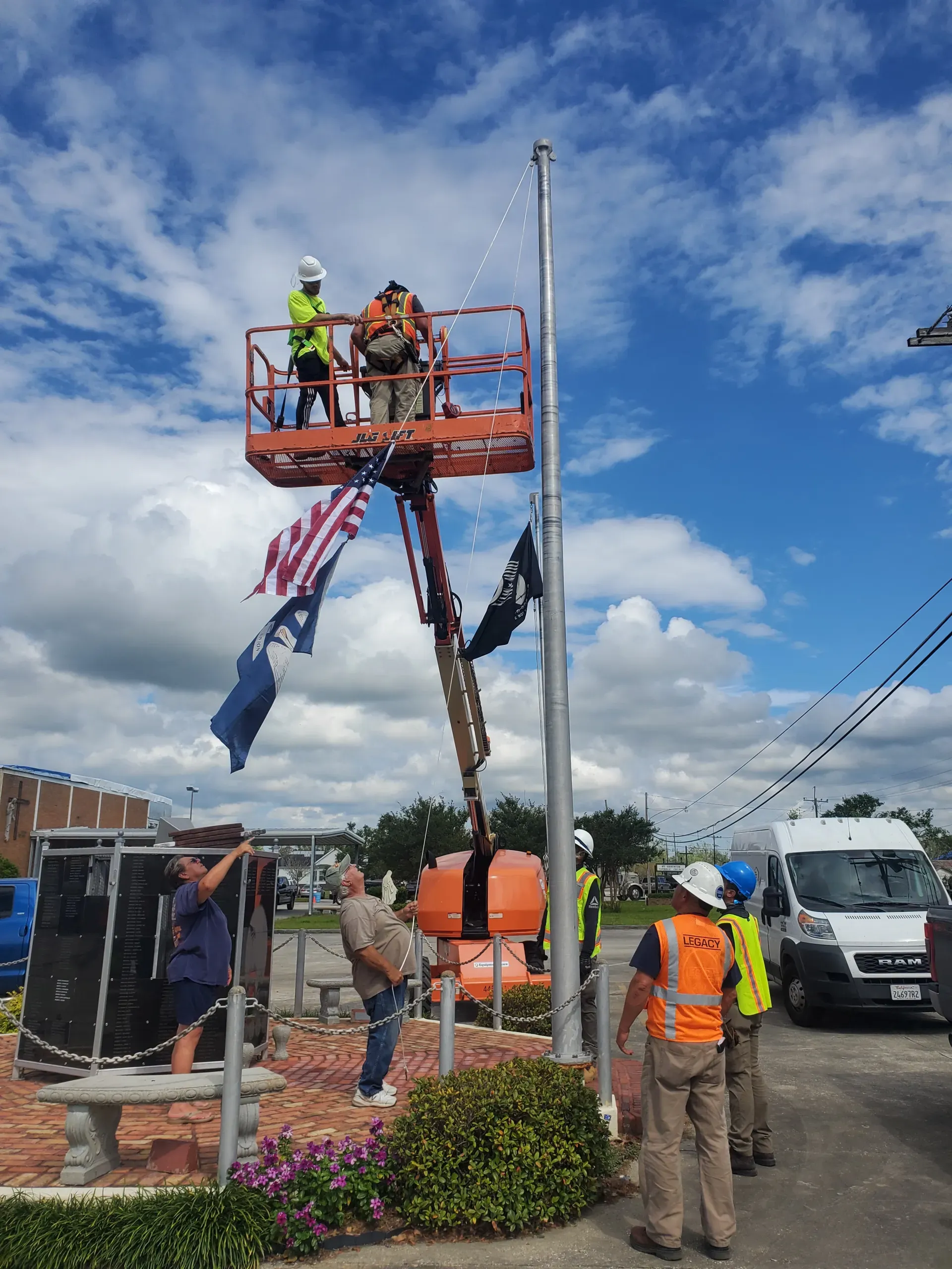 Utility workers on a lift fixing a pole beside a road, with trucks, cones, and flags under a blue sky