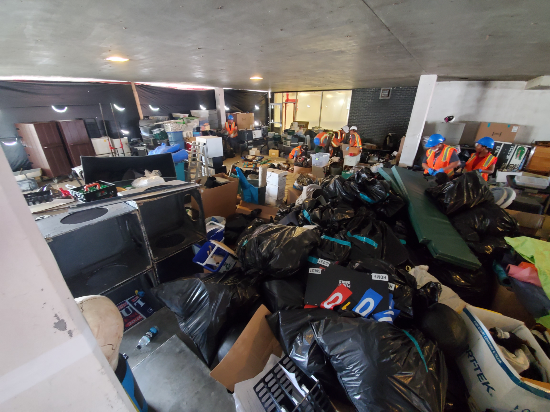 Cluttered indoor storage area packed with bags, boxes, and assorted piled items under fluorescent lights