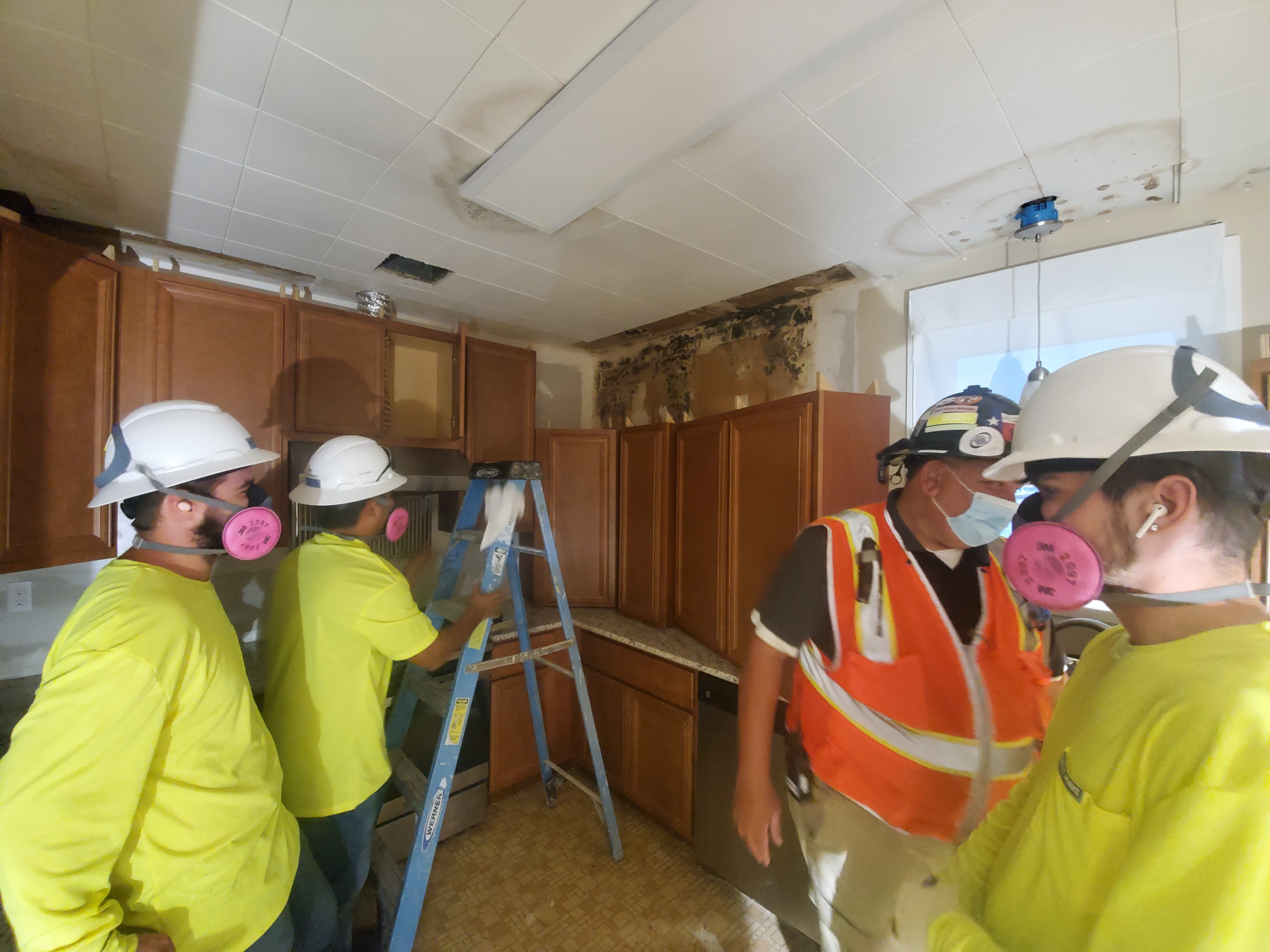 Workers in hard hats inspect a damaged room with exposed ceiling, debris, and wooden cabinets.