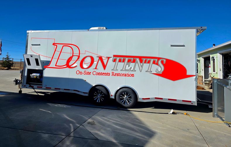 White Deon Tents trailer parked outdoors under a blue sky, with red logo and lettering.