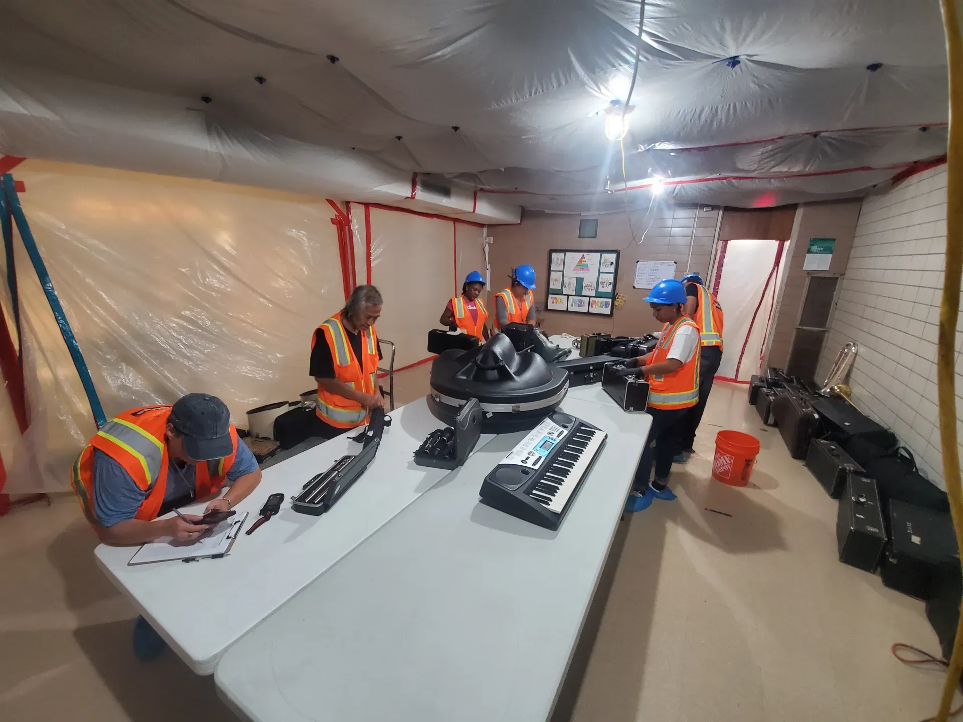 Workers in orange safety vests and blue hard hats sort gear inside a temporary tent workspace.